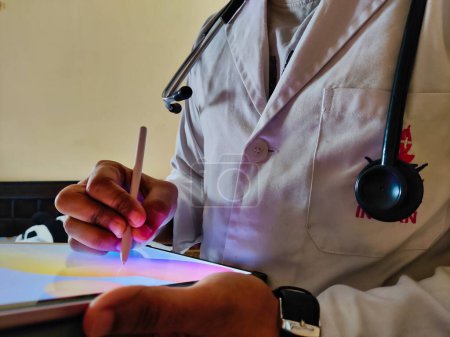 Téléchargez les photos : Picture of a doctor wearing a white apron and stethoscope using a tablet. Writing prescription, searching for Neet PG, NEXT exam. - en image libre de droit