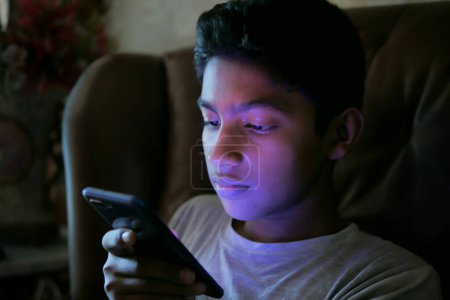 Téléchargez les photos : Adolescent garçon assis sur canapé en utilisant un téléphone intelligent la nuit . - en image libre de droit