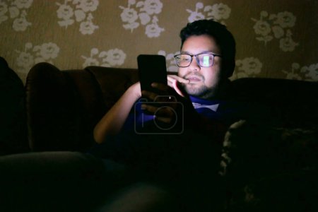 Téléchargez les photos : Jeune homme assis sur le lit en utilisant un téléphone intelligent la nuit . - en image libre de droit