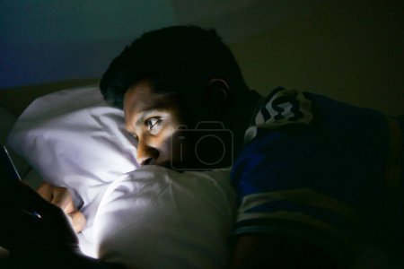 Téléchargez les photos : Jeune homme assis sur le lit en utilisant un téléphone intelligent la nuit . - en image libre de droit