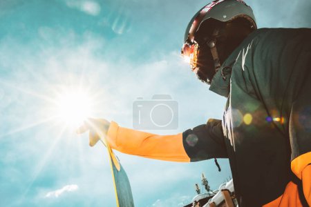 Photo for Close up portrait snowboarder stand hold board look up to sky to sun black copyspace with sunburst in sunny day. Ski holidays - Royalty Free Image