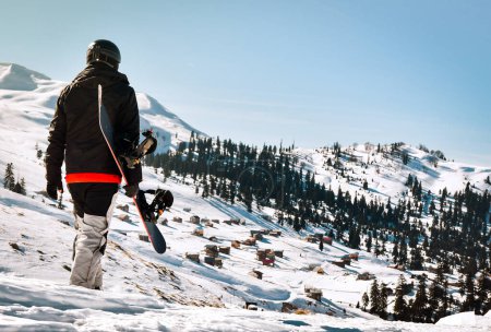 Téléchargez les photos : Homme touriste en combinaison de ski avec snowboard dans ses mains promenades sur la pente enneigée dans le contexte de beau paysage de montagne - en image libre de droit