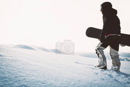 Téléchargez les photos : Snowboarder marche avec snowboard pendant le coucher du soleil dans les montagnes enneigées. Freerider solo cinématique fond snowboarder - en image libre de droit