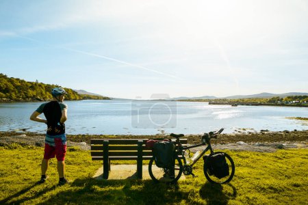 Photo for Male cyclist stand by bicycle rest in scenic nature by lake relax enjoy evening in summer. Ireland cycle outdoors.Eco green travel - Royalty Free Image