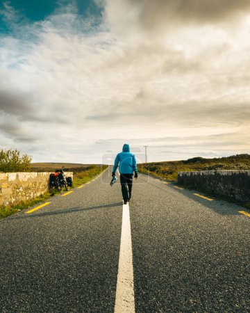 Photo for Back view male cyclist in blue jacket walk on road by touring bicycle travel outdoors in Ireland. Purpose and travel adventure concept. Wild atlantic way road trip - Royalty Free Image