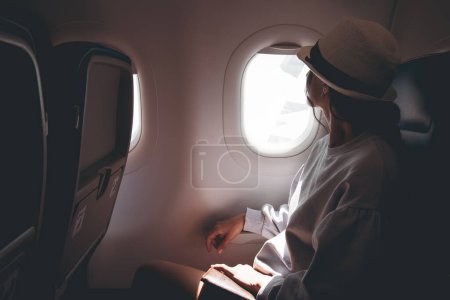 Woman looks out the window of an flying airplane. Young caucasian happy passengers are traveling by plane, watching the sky from above and hold passport