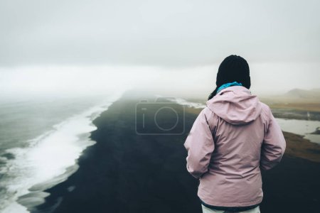 Mujer turista de pie mirada reflexiva a las olas del océano atlántico. Famoso mirador icónico acantilado sobre la playa de arena negra de Reynisfjara. Persona busca dirección y propósito en los viajes