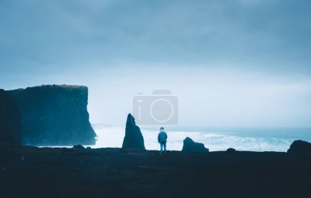 Woman tourist stand in very windy day watch Atlantic ocean waves crash to rocks in Reykjanes peninsula shore in Iceland. Popular scenic nature location. Iconic Valahnukamol stack