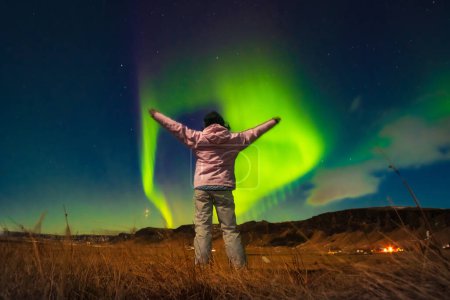 Woman watches the northern lights arms up excited joyful in Iceland. Woman in Icelandic spring night landscape. Iceland travel chasing northern lights concept