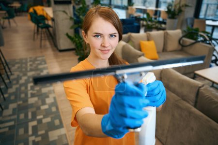 Photo for Red-haired cleaning lady in uniform and protective gloves uses glass scraper and spray to work - Royalty Free Image