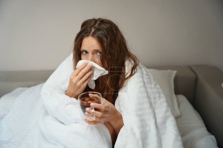 Photo for Sick lady sits in bed wrapped in blanket while she has cup of hot tea in hand - Royalty Free Image