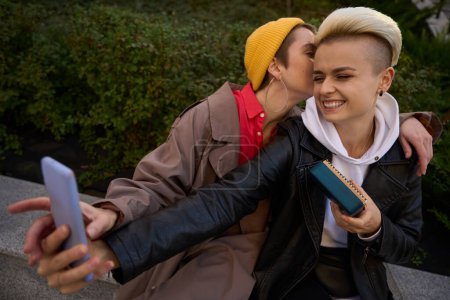 Téléchargez les photos : Blonde prend un selfie avec sa copine sur fond de buisson pittoresque, la brune murmure à l'oreille - en image libre de droit