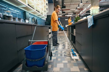 Photo for Young woman cleaner washes the floor with a mop, next to a cart with buckets - Royalty Free Image