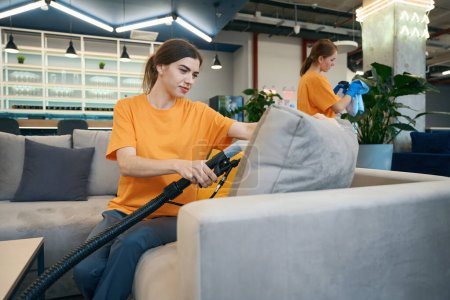 Photo for Cleaners cleaning company perform thorough cleaning furniture with a vacuum cleaner in coworking space and take care of the plants - Royalty Free Image