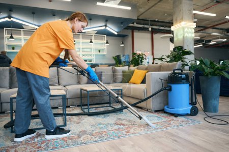 Femme de l'entreprise de nettoyage dans un uniforme confortable nettoie le tapis avec un aspirateur professionnel dans un espace de coworking