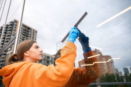 Photo for Cleaning service worker in a warm sweatshirt washes the mirrored windows of the office center, uses a glass scraper - Royalty Free Image