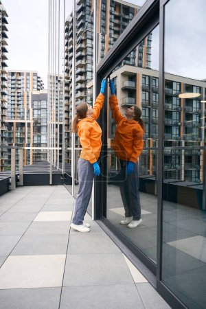Photo for Employee cleaning service washes mirrored windows of the office center with a glass scraper, she is in uniform and gloves - Royalty Free Image