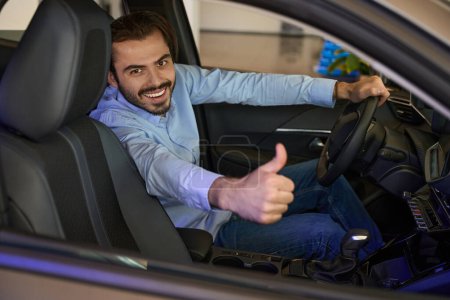 Foto de Sonriente conductor de coche alegre sentado detrás del volante y haciendo pulgares hacia arriba signo - Imagen libre de derechos