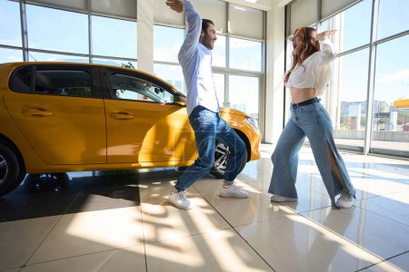 Téléchargez les photos : Joyeux élégant jeune acheteur de véhicules et son compagnon dansant dans le showroom automobile - en image libre de droit