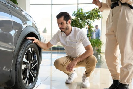 Photo for Focused man sitting on his haunches and inspecting automobile tire condition in presence of sales consultant - Royalty Free Image