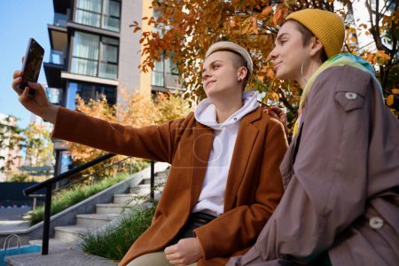 Téléchargez les photos : Couple féminin Lgbt prendre un selfie dans la ville d'automne sur fond de grands bâtiments - en image libre de droit