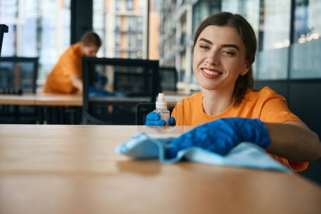 Photo for Smiling female cleaners are cleaning in the office with detergents, they are wearing yellow clothes and blue gloves - Royalty Free Image