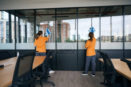 Photo for Two female employees of a cleaning company washing windows in a coworking space with a special product in protective gloves - Royalty Free Image