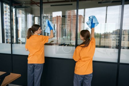 Téléchargez les photos : Les femmes employées de l'entreprise de nettoyage lavent les fenêtres dans l'espace de coworking, elles sont habillées en salopette et travaillent avec des gants de protection - en image libre de droit