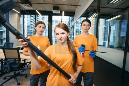 Photo for Three workers cleaning company in uniform hold working gadgets in their hands - vacuum cleaner, spray gun and special spray - Royalty Free Image