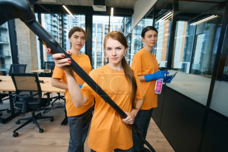 Photo for Three cleaning company workers in uniform and protective gloves cleaning and disinfecting office space with vacuum cleaner, rag and spray - Royalty Free Image