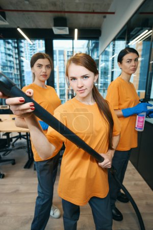 Photo for Three female workers cleaning company in protective gloves cleaning office space with vacuum cleaner, a rag and a spray bottle - Royalty Free Image