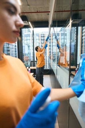 Foto de Los empleados de la empresa de limpieza en overoles tratan tabiques de vidrio en el espacio de coworking con un desinfectante, trabajan en guantes de goma - Imagen libre de derechos