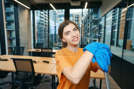 Téléchargez les photos : Jolie femme de ménage souriante dans un espace de coworking dans des moments de détente, elle est en uniforme et gants en caoutchouc - en image libre de droit