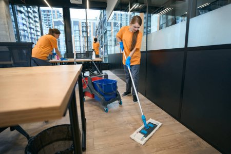 Foto de Equipo de limpiadores trabajan en un área de coworking, una mujer lava el suelo, ventanas t muebles - Imagen libre de derechos