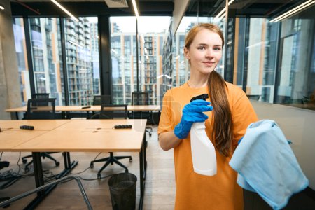 Téléchargez les photos : Femme de ménage dans la zone de coworking avec vaporisateur et serviette dans ses mains, elle est en uniforme jaune et gants bleus - en image libre de droit