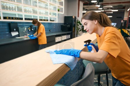 Téléchargez les photos : Les femmes nettoyantes dans la zone de coworking nettoient et désinfectent les surfaces, elles utilisent un vaporisateur et des lingettes - en image libre de droit