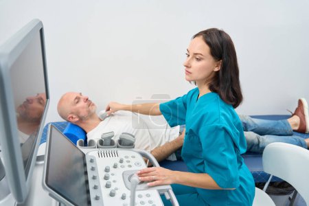 Foto de Médico serio en uniforme médico haciendo ecografía tiroidea de un hombre mientras está acostado en un sofá - Imagen libre de derechos