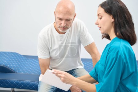 Photo for Enthusiastic woman in medical uniform consulting a man while he carefully looks at the printout in her hands - Royalty Free Image