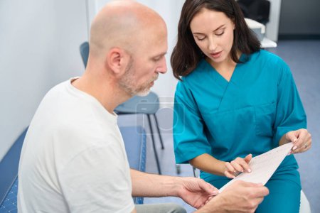 Foto de Mujer enfocada en uniforme médico consultando a un hombre mientras él mira cuidadosamente la impresión en sus manos - Imagen libre de derechos