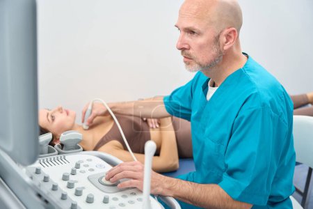 Téléchargez les photos : Homme concentré dans le bureau médical pointant un dispositif d'examen d'organe interne au cou du patient alors qu'elle est allongée sur le canapé - en image libre de droit