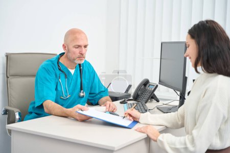 Photo for Serious doctor in medical uniform holding a clipboard in his hands while the patient in front puts his signature on it - Royalty Free Image