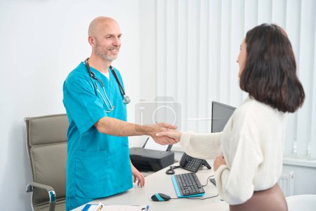 Téléchargez les photos : Satisfait médecin en uniforme médical accueille femme alors qu'ils se trouvent en face de l'autre dans son cabinet - en image libre de droit