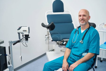 Foto de Ginecólogo sonriente está sentado en un consultorio médico mientras lleva un uniforme y un estetoscopio cuelga alrededor de su cuello - Imagen libre de derechos