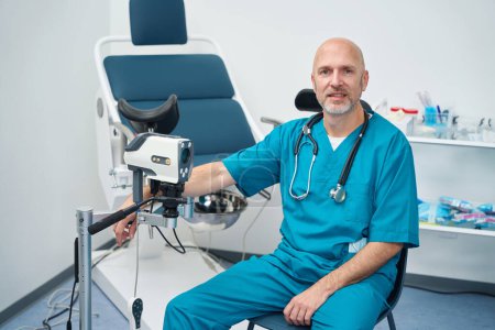 Téléchargez les photos : Gynécologue sérieux est assis dans un cabinet médical tandis que sa main reposait sur un détail d'une chaise pour les manipulations gynécologiques - en image libre de droit