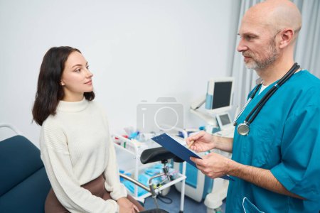 Photo for Calm female sits on a chair in front of a medical officer while he holds a folder and looks at her - Royalty Free Image