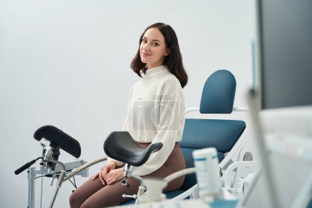 Téléchargez les photos : Joyeuse patiente tenant un bilan de santé féminin en attendant un médecin assis sur une chaise gynécologique - en image libre de droit