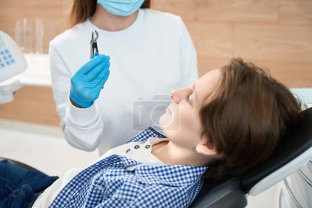 Téléchargez les photos : Garçon regarde outil spécial pour l'extraction des dents dans les mains d'un dentiste, un médecin en gants et un masque - en image libre de droit