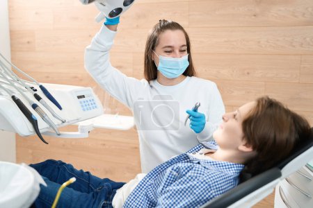 Téléchargez les photos : Femme dentiste amicale encourage une jeune patiente avant l'extraction des dents, elle utilise des outils spéciaux - en image libre de droit