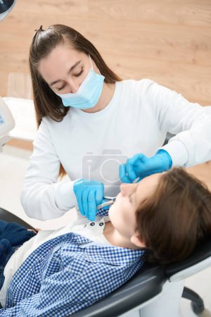 Téléchargez les photos : Chirurgien dentiste enlève une dent à un garçon avec un outil spécial, une femme travaille dans une pièce lumineuse - en image libre de droit