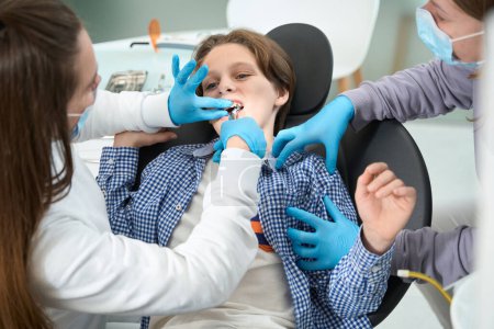 Téléchargez les photos : Dentiste-chirurgien enlève une dent à un adolescent avec un outil spécial, un assistant tient le garçon - en image libre de droit
